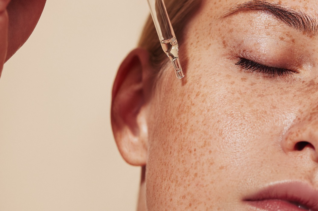 Nahaufnahme mit hoher Detailgenauigkeit von weiblicher Haut mit Sommersprossen und einer Pipette mit Serum. Ausschnitt einer jungen Frau mit glatter, makelloser Haut, die flüssiges Serum auf ihr Gesicht aufträgt.